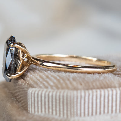 Close-up side view of an elegant gold ring set with a large oval salt and pepper diamond. It features tiger-style prongs atop a decorative under basket, displayed on a textured beige fabric surface.