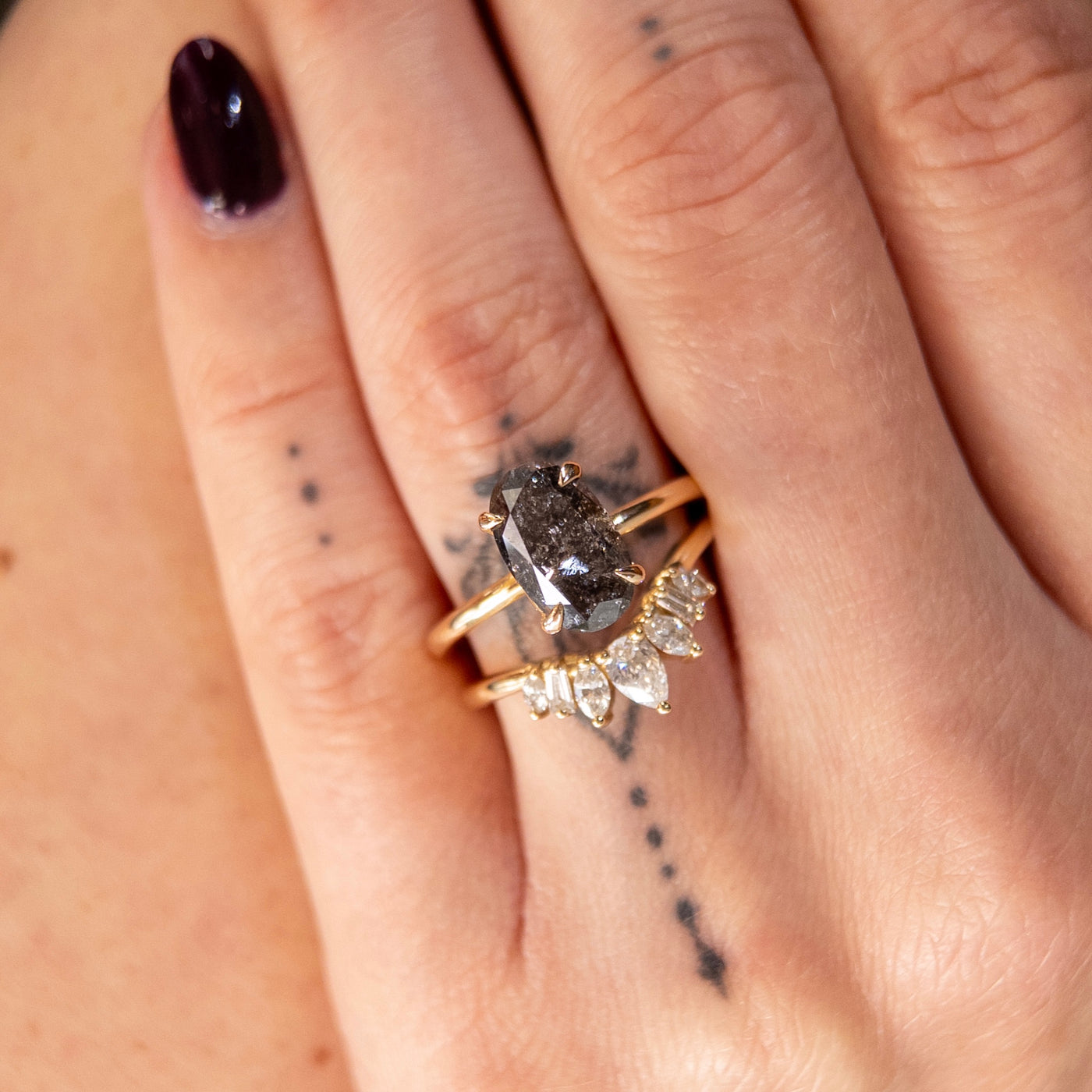 An elegant gold ring set with a large oval salt and pepper diamond. It features tiger-style prongs atop a decorative under basket, paired with a multi-shaped diamond band. Worn on a hand. 