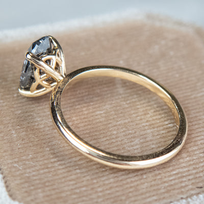 Close-up side view of an elegant gold ring set with a large oval salt and pepper diamond. It features tiger-style prongs atop a decorative under basket, displayed on a textured beige fabric surface.