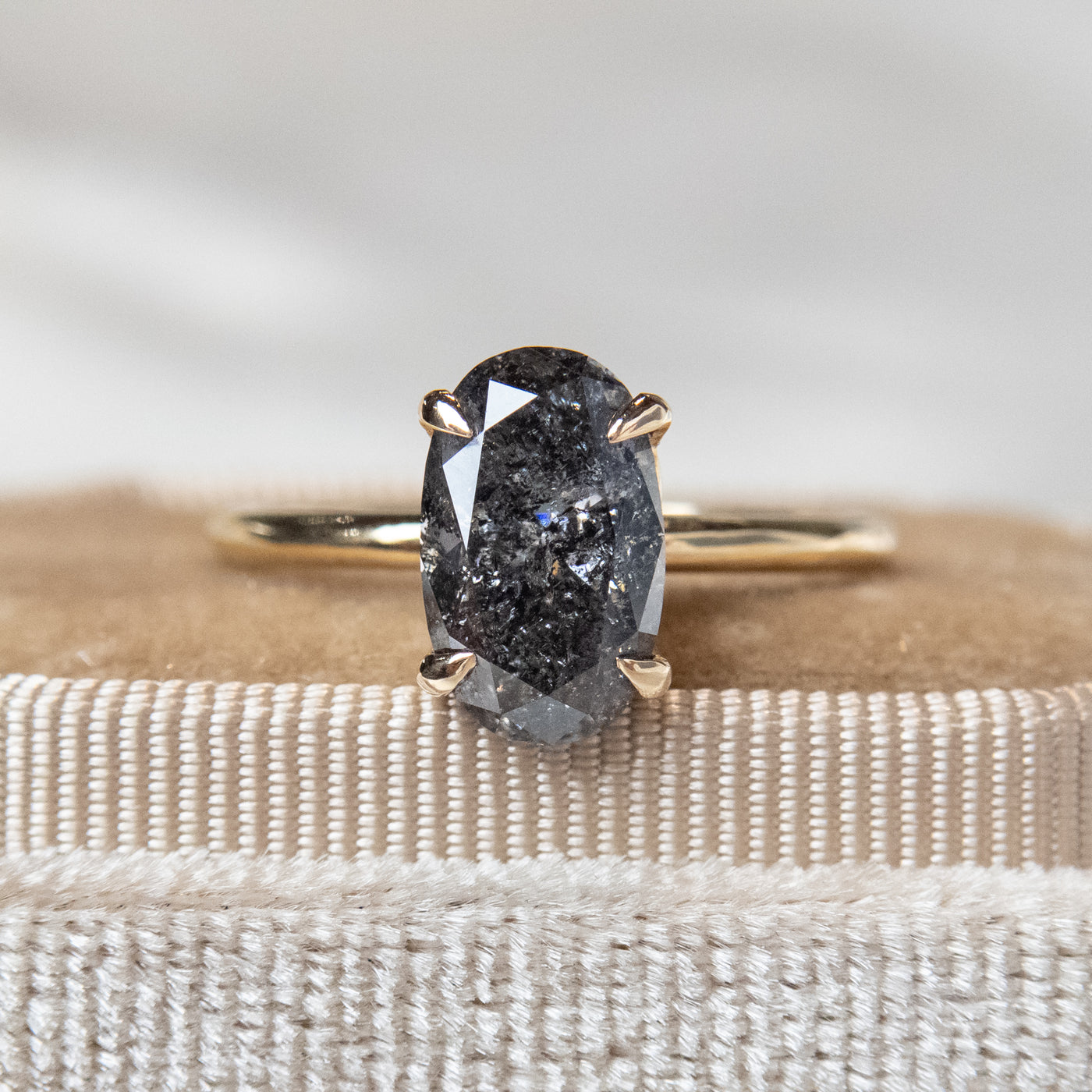 Close-up of an elegant gold solitaire ring set with a large oval salt and pepper diamond, placed on a textured beige fabric surface.