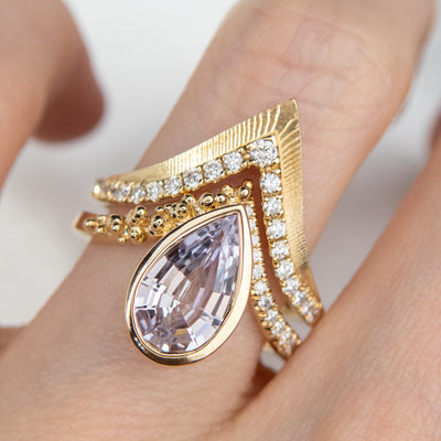 Elegant gold ring with a V-shaped design, encrusted with sparkling diamonds along the band stacked on a finger with a geometric purple pear shaped sapphire engagement ring.