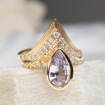 Elegant gold ring with a V-shaped design, encrusted with sparkling diamonds along the band stacked with a geometric purple pear shaped sapphire engagement ring.