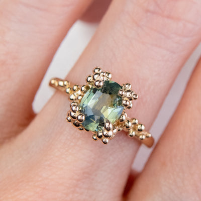 A gold ring with a cluster of small round gold beads holds an oval green sapphire at the center, worn on a finger.  