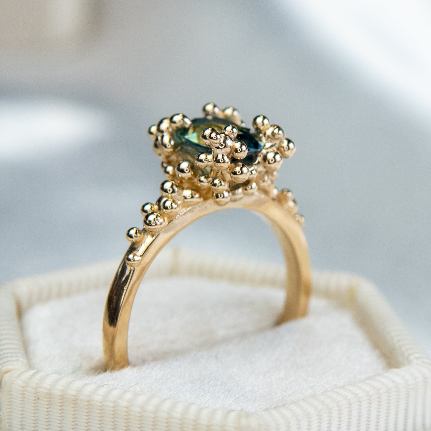 Side view of a gold ring with a cluster of small round gold beads holds an oval green sapphire at the center, displayed in a cream ring box.