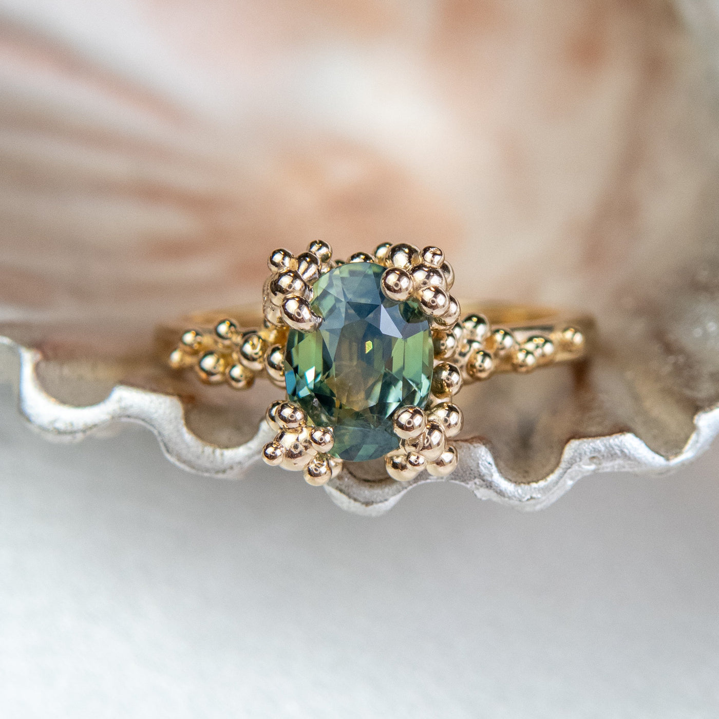 A gold ring with a cluster of small round gold beads holds an oval green sapphire at the center, displayed on a metallic textured surface.