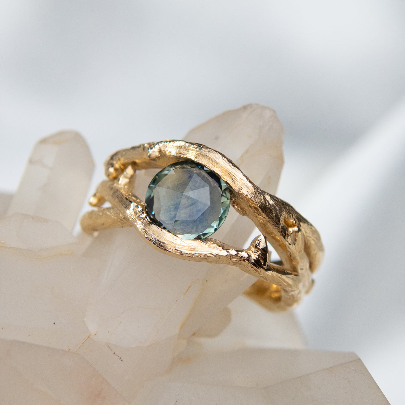 Gold ring with a unique twisted band design, featuring a round rose cut teal blue sapphire, displayed on a white crystal background.