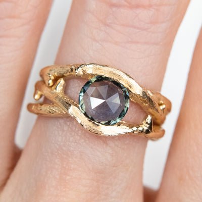 Gold ring with a unique twisted band design, featuring a round rose cut teal blue sapphire, worn on a a finger.