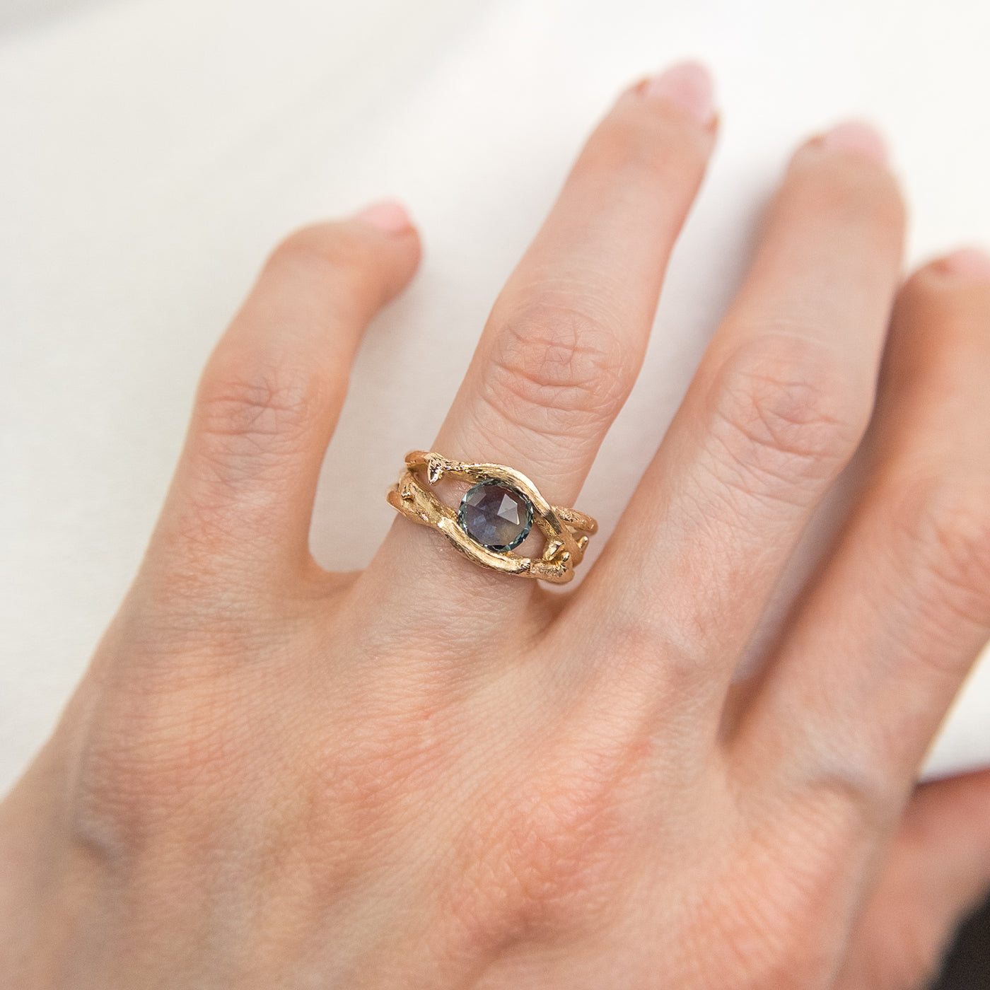 Gold ring with a unique twisted band design, featuring a round rose cut teal blue sapphire, worn on a a hand.