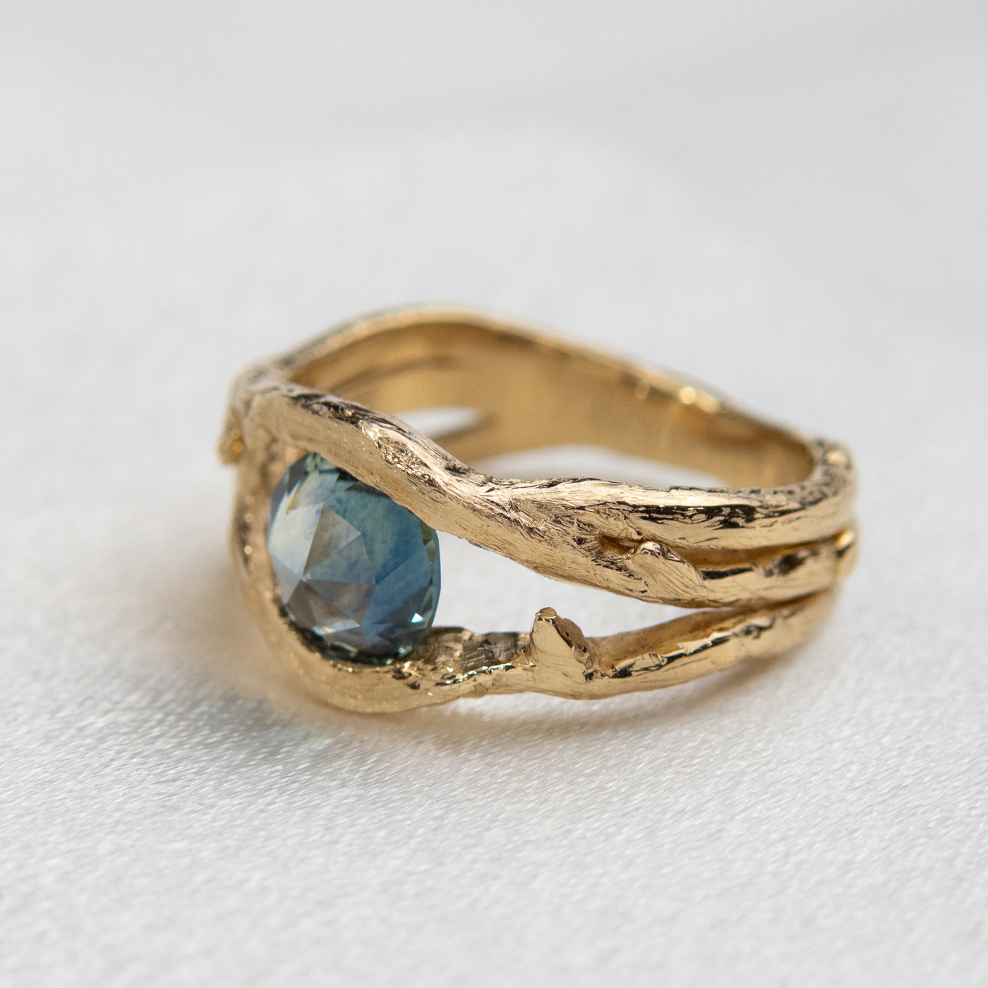 Side angle view of a gold ring with a unique twisted band design, featuring a round rose cut teal blue sapphire, displayed on a white crystal background.