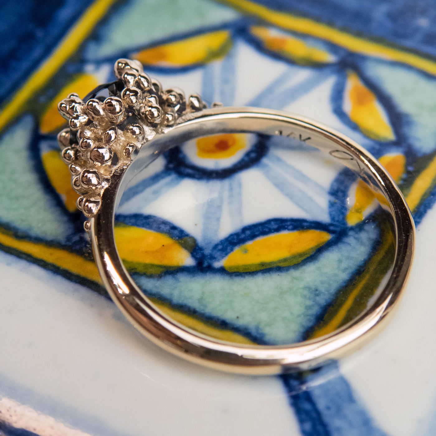 A profile view of a gold ring with a textured band that features a round brilliant cut blue sapphire, set in ornate gold prongs, set atop a colourful tile. 