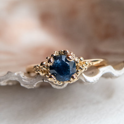 A gold ring with a textured band features a round brilliant cut deep blue sapphire, set in ornate gold prongs. 