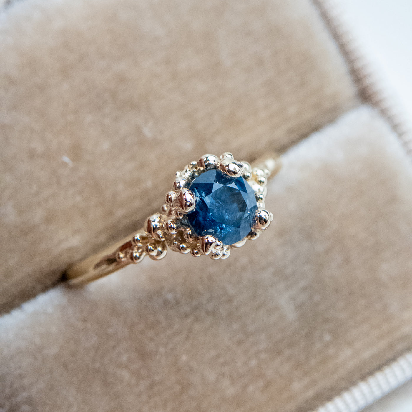 A gold ring with a textured band features a round brilliant cut blue sapphire, set in ornate gold prongs, in a soft fabric ring box. 