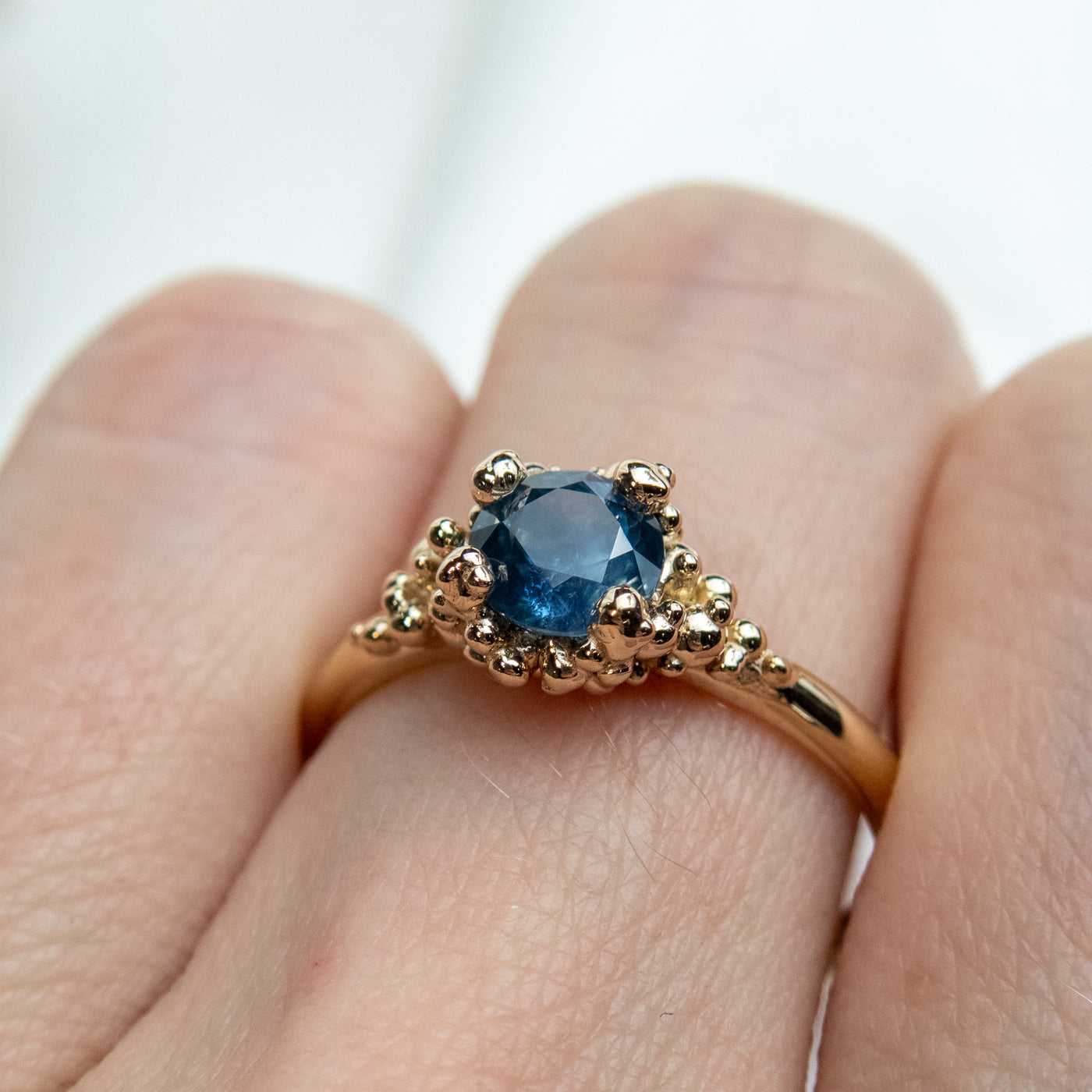 A gold ring with a textured band features a round brilliant cut blue sapphire, set in ornate gold prongs, worn on a finger 