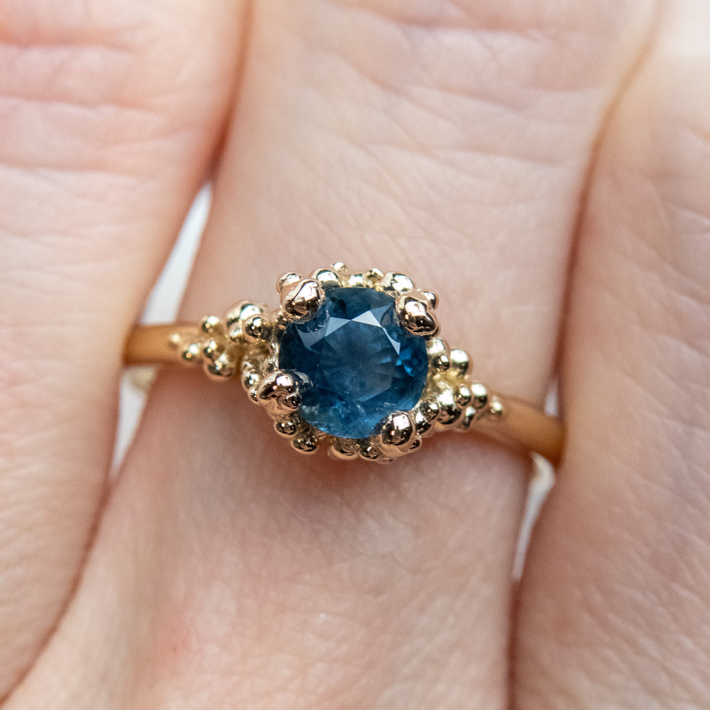 A gold ring with a textured band features a round brilliant cut blue sapphire, set in ornate gold prongs, worn on a finger 