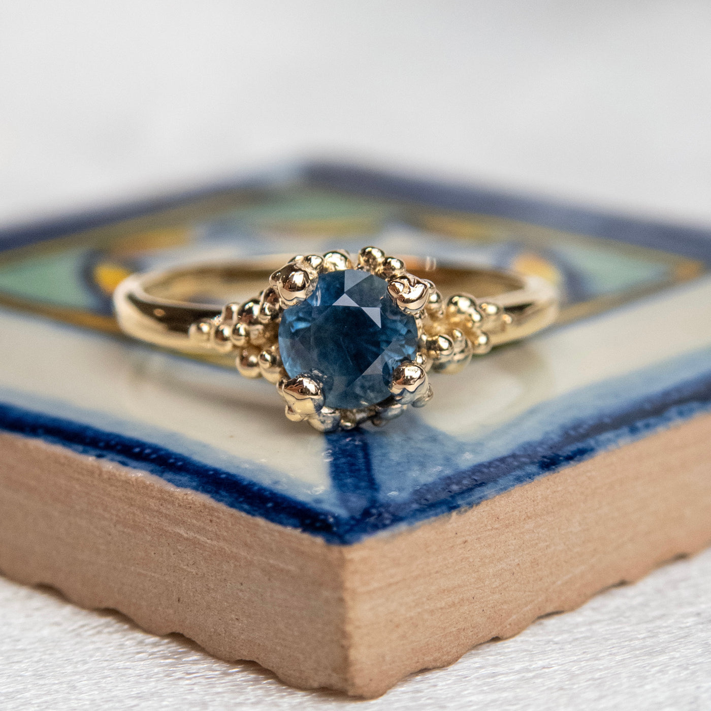 A gold ring with a textured band features a round brilliant cut blue sapphire, set in ornate gold prongs, set atop a colourful tile. 