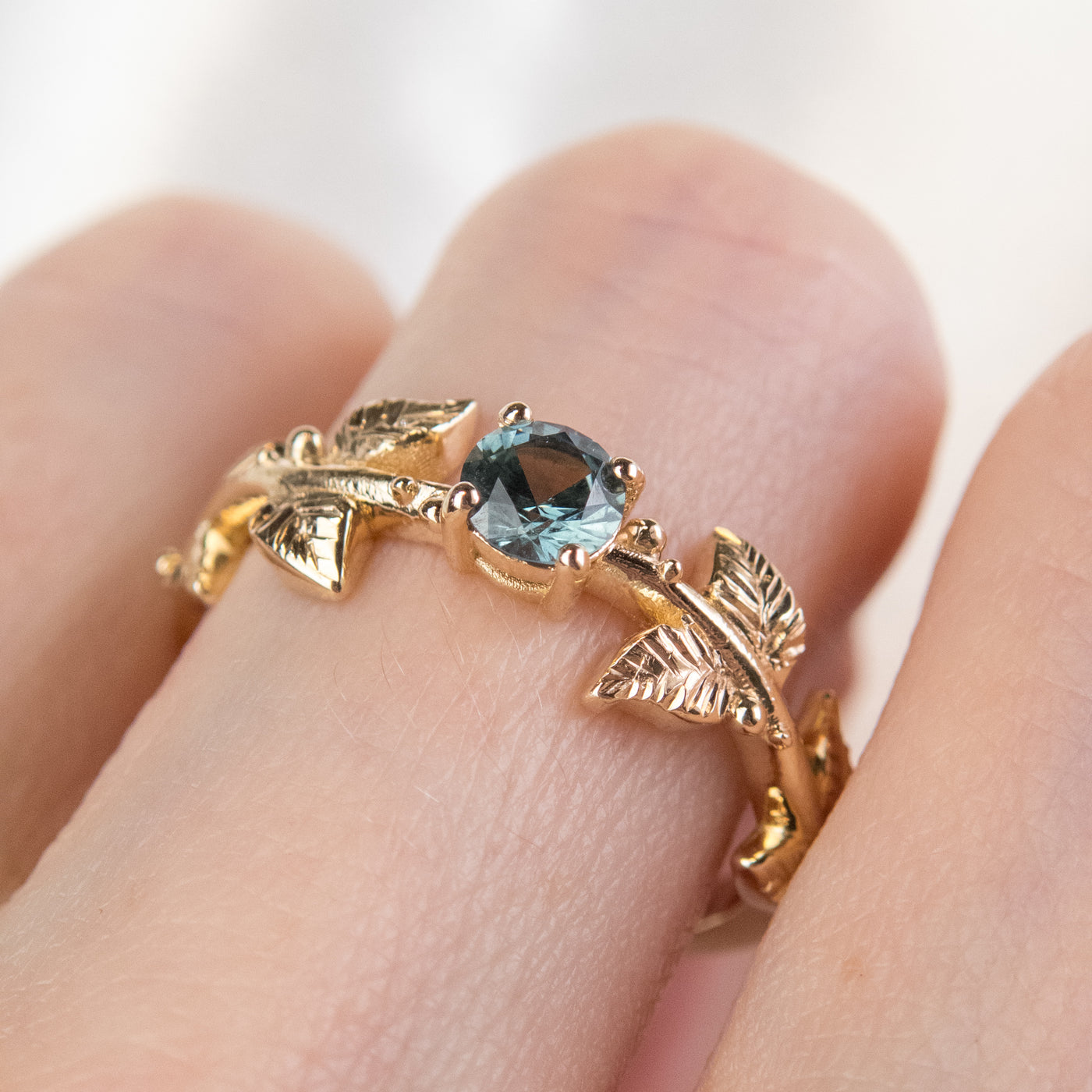 Nature-inspired gold ring with a round brilliant teal sapphire, featuring intricate leaf design band. Worn on a finger. 