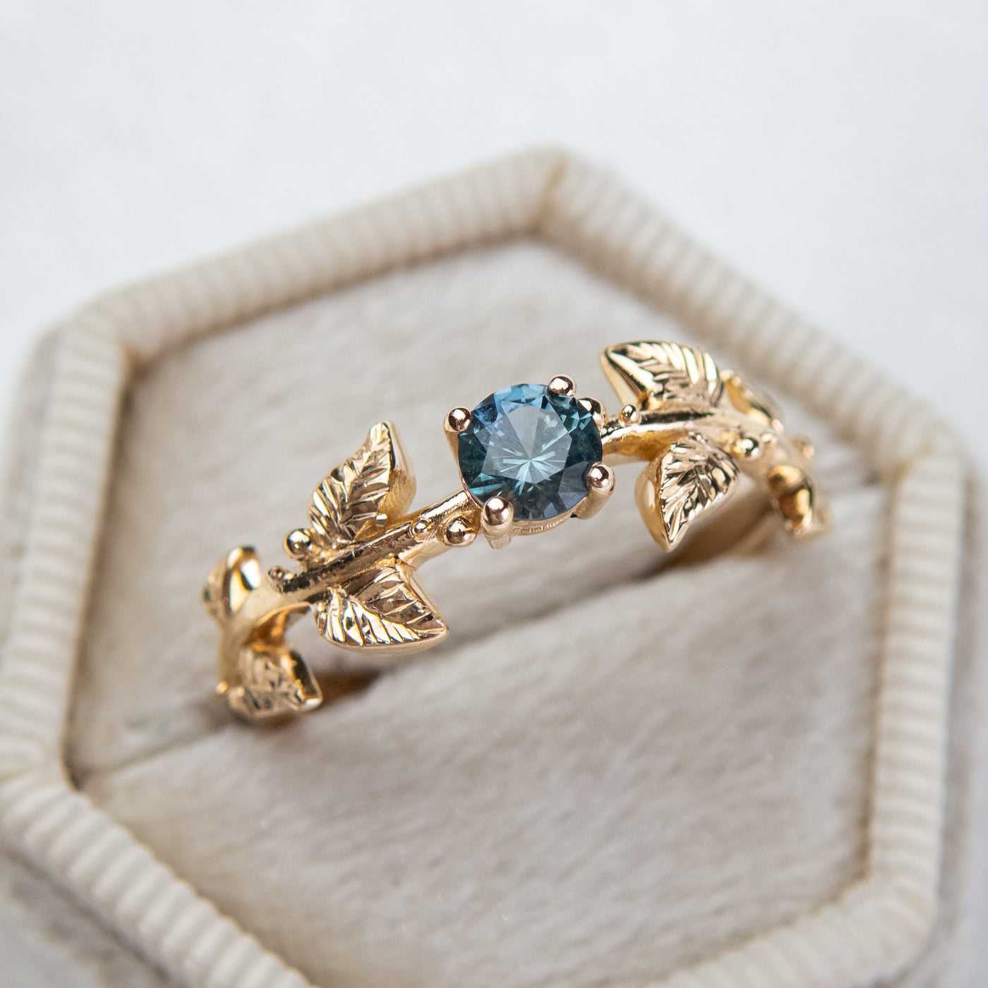 Nature-inspired gold ring with a round brilliant teal sapphire, featuring intricate leaf design band. Set in a light-colored, hexagonal velvet box.