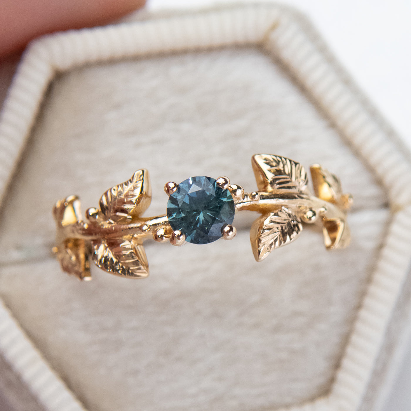 Nature-inspired gold ring with a round brilliant teal sapphire, featuring intricate leaf design band. Set in a light-colored, hexagonal velvet box.