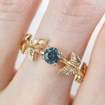 Nature-inspired gold ring with a round brilliant teal sapphire, featuring intricate leaf design band. Worn on a finger. 