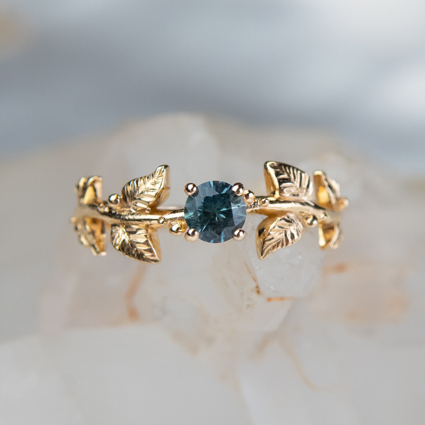 Nature-inspired gold ring with a round brilliant teal sapphire, featuring intricate leaf design band. Set against a light crystalline background. 