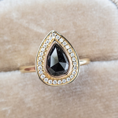 A pear-shaped black diamond ring is set in gold, surrounded by a halo of small, sparkling white diamonds. Displayed in a beige ring box. 
