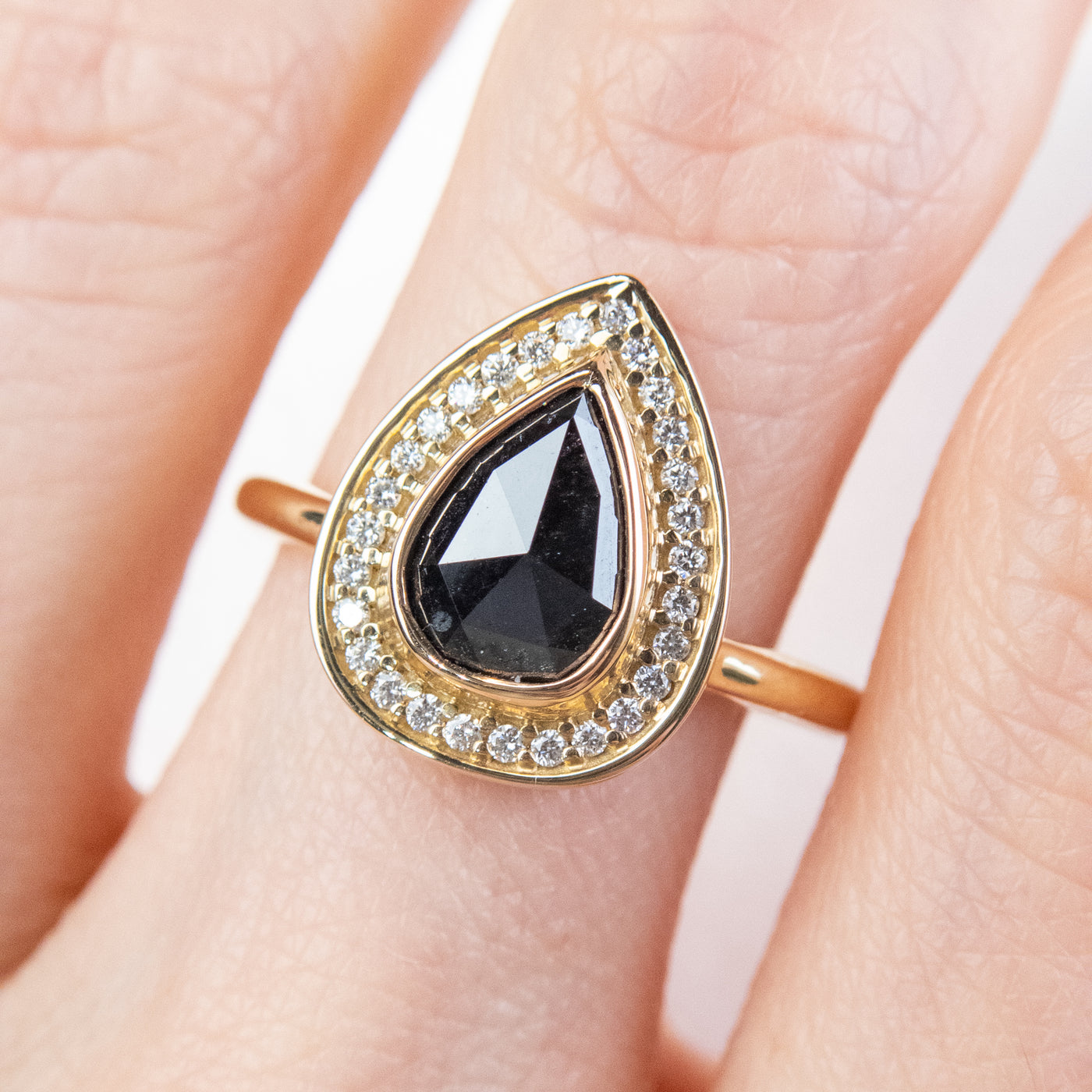 A pear-shaped black diamond ring is set in gold, surrounded by a halo of small, sparkling white diamonds. Worn on a finger. 