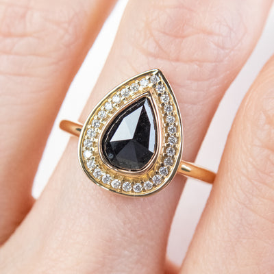 A pear-shaped black diamond ring is set in gold, surrounded by a halo of small, sparkling white diamonds. Worn on a finger. 