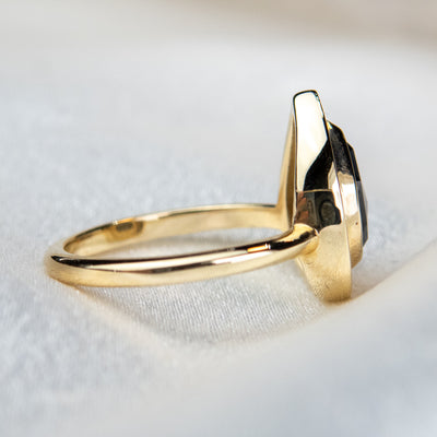 A side view of a pear-shaped black diamond ring is set in gold, surrounded by a halo of small, sparkling white diamonds. The background is a soft beige fabric.