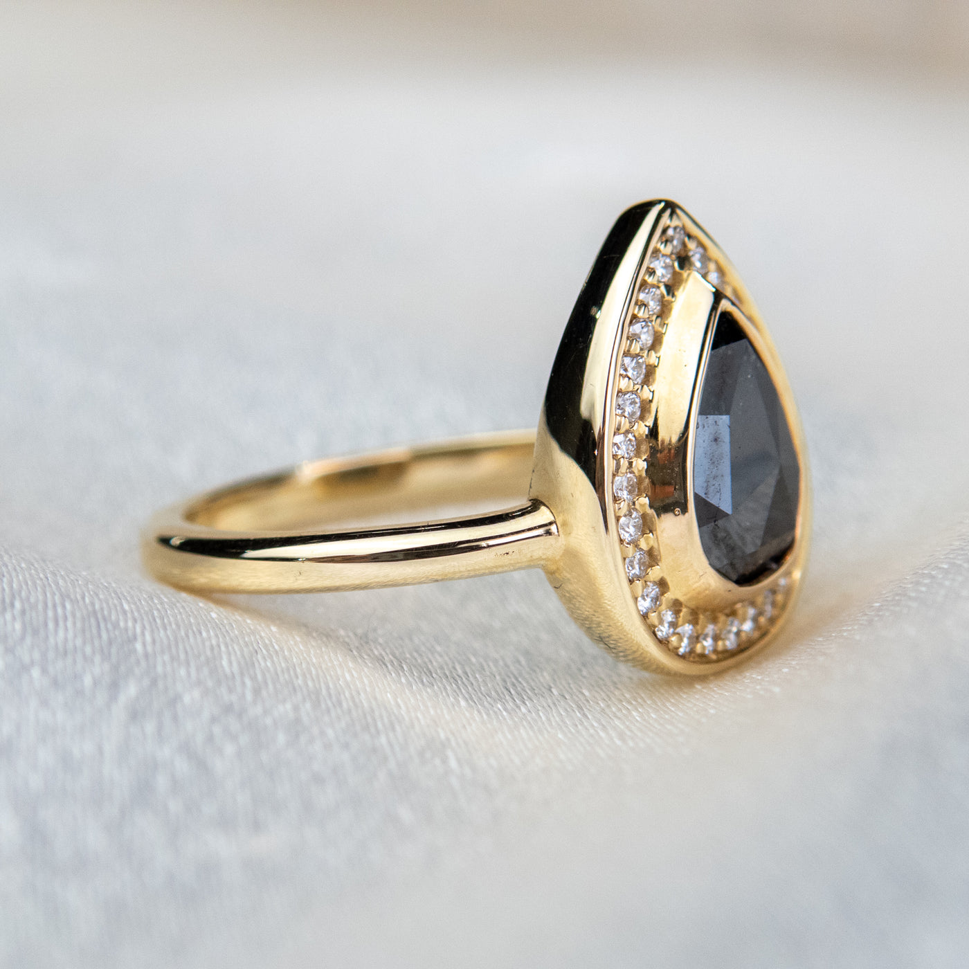 A side view of a pear-shaped black diamond ring is set in gold, surrounded by a halo of small, sparkling white diamonds. The background is a soft beige fabric.