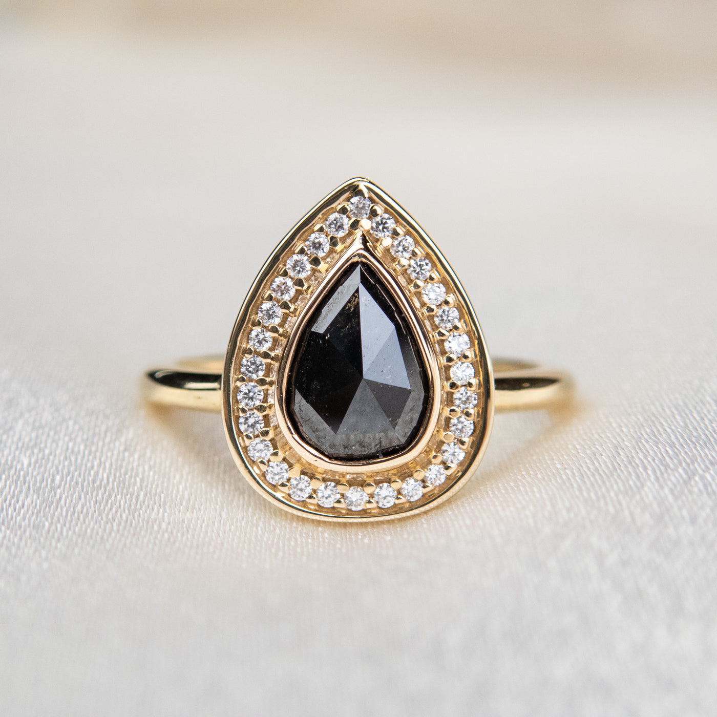 A pear-shaped rose cut black diamond ring is set in gold, surrounded by a halo of small, sparkling white diamonds. The background is a soft beige fabric.