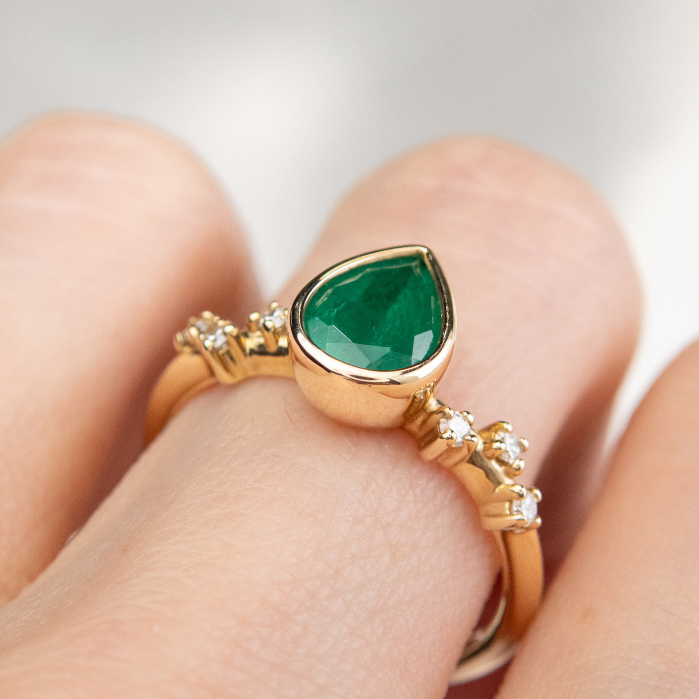 A pear-shaped emerald set in a gold bezel ring, adorned with offset small diamonds on the band. worn on a hand. 