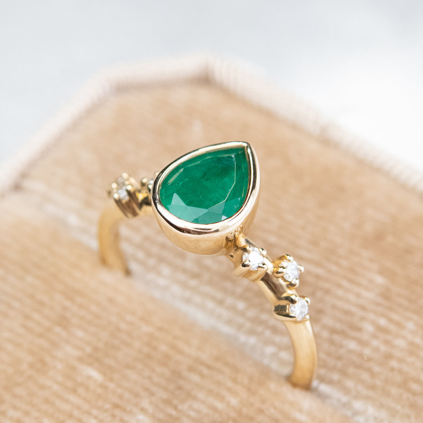 A pear-shaped emerald set in a gold bezel ring, adorned with offset small diamonds on the band. Displayed in a beige velvet ring box.