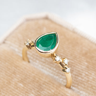 A pear-shaped emerald set in a gold bezel ring, adorned with offset small diamonds on the band. Displayed in a beige velvet ring box.