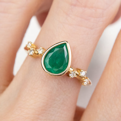 A pear-shaped emerald set in a gold bezel ring, adorned with offset small diamonds on the band. worn on a hand. 