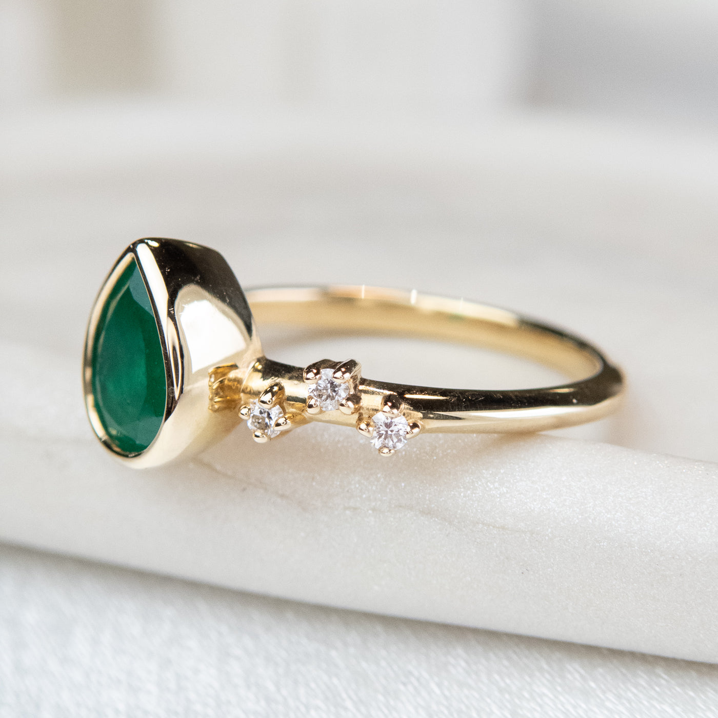 A pear-shaped emerald set in a gold bezel ring, adorned with offset small diamonds on the band. Displayed on a light background. 