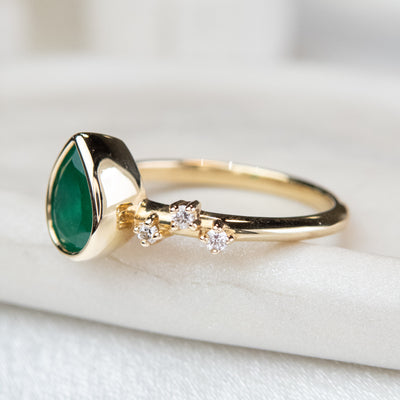 A pear-shaped emerald set in a gold bezel ring, adorned with offset small diamonds on the band. Displayed on a light background. 