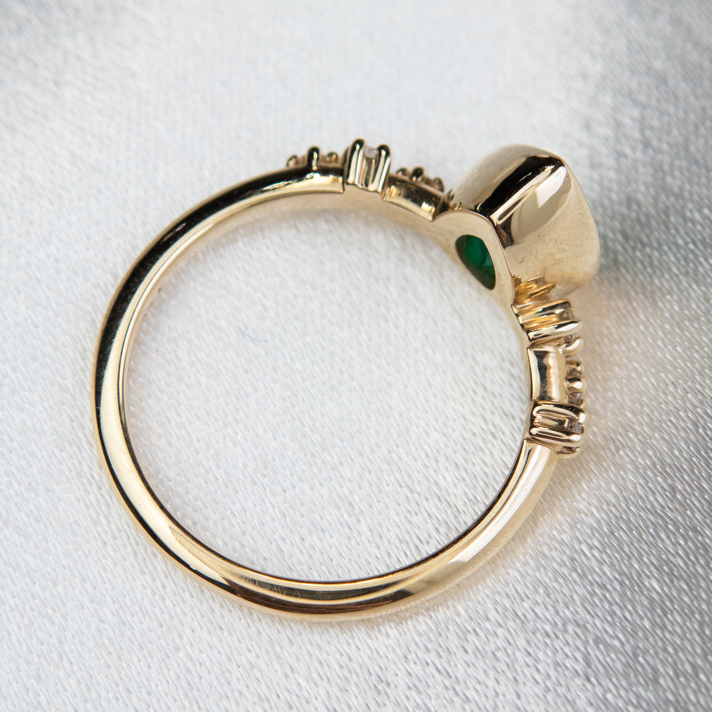 A top view of a pear-shaped emerald set in a gold bezel ring, adorned with offset small diamonds on the band. Displayed on a light background. 