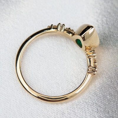 A top view of a pear-shaped emerald set in a gold bezel ring, adorned with offset small diamonds on the band. Displayed on a light background. 