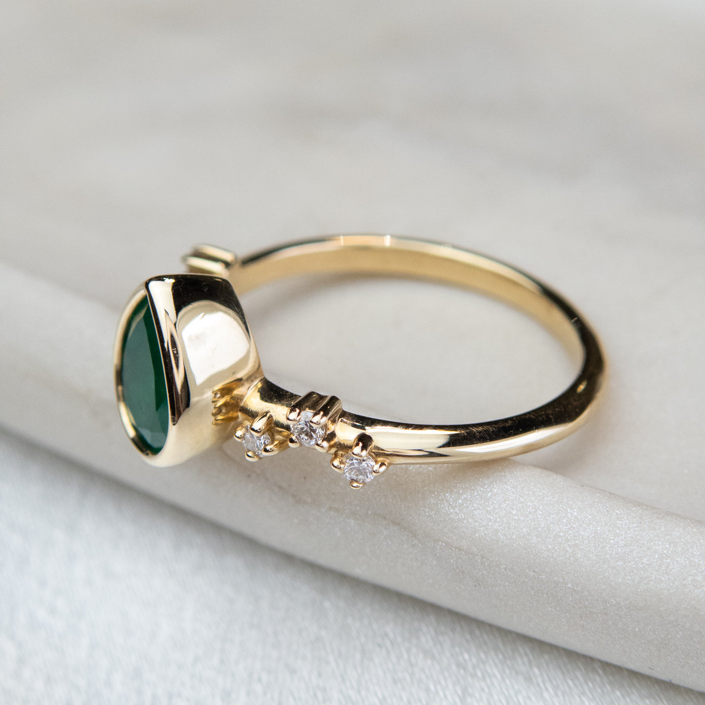 A pear-shaped emerald set in a gold bezel ring, adorned with offset small diamonds on the band. Displayed on a light background. 