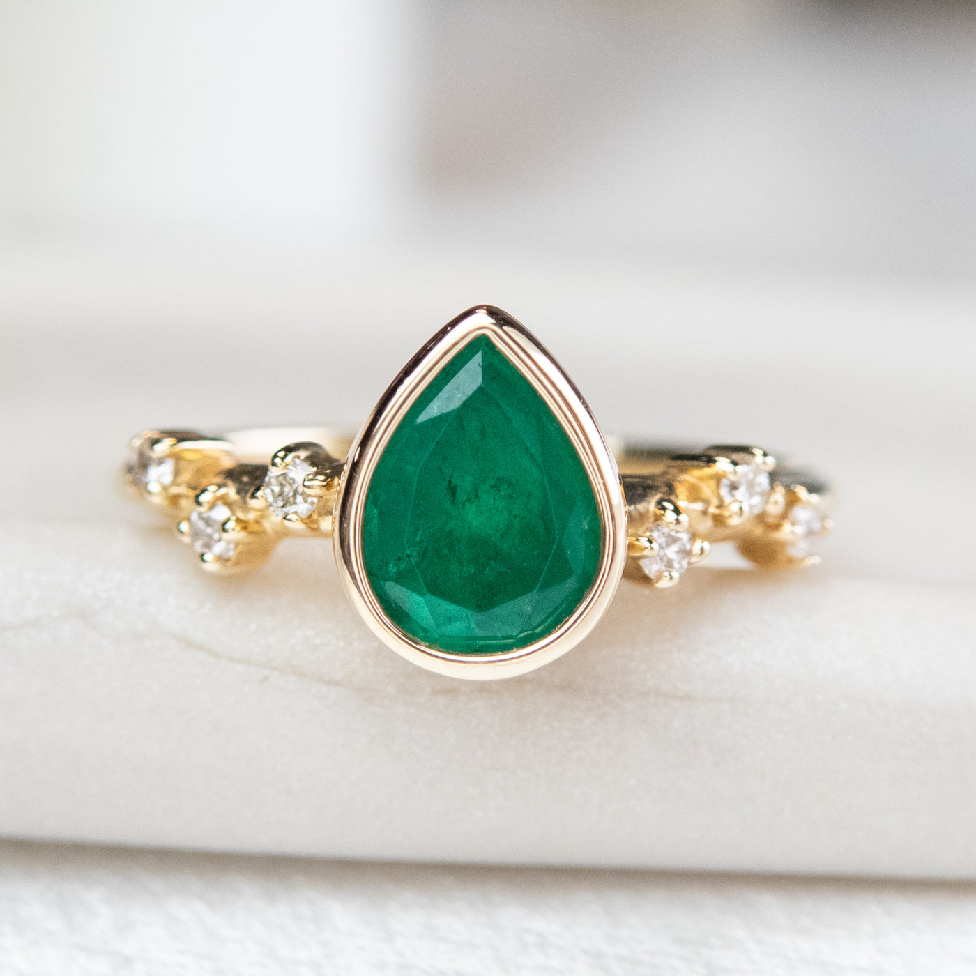 A pear-shaped emerald set in a gold bezel ring, adorned with offset small diamonds on the band. Displayed on a light background. 