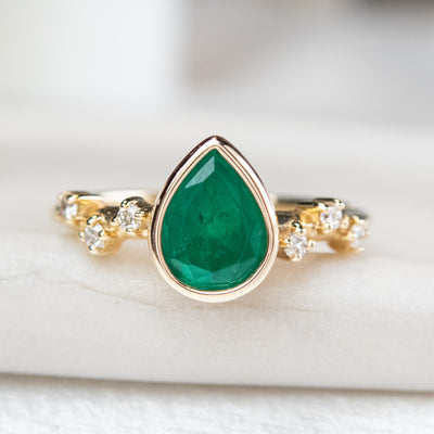 A pear-shaped emerald set in a gold bezel ring, adorned with offset small diamonds on the band. Displayed on a light background. 
