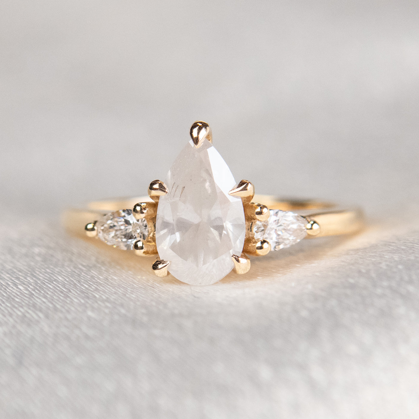 A three stone ring with a central pear-shaped diamond set in gold , flanked by two smaller pear diamonds, reflecting light on a soft, light, blurred background. 