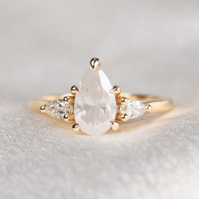 A three stone ring with a central pear-shaped diamond set in gold , flanked by two smaller pear diamonds, reflecting light on a soft, light, blurred background. 