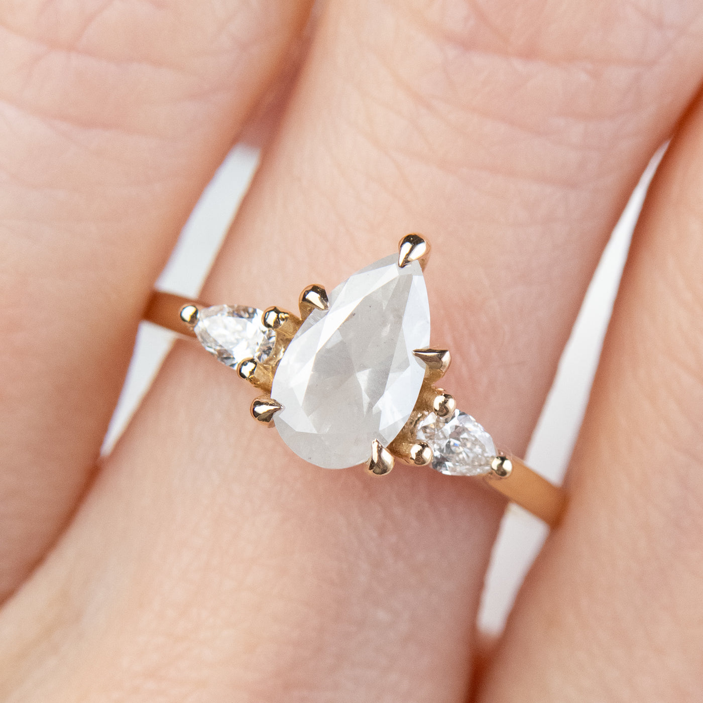 A three stone ring with a central pear-shaped diamond set in gold , flanked by two smaller pear diamonds, worn on a hand. 