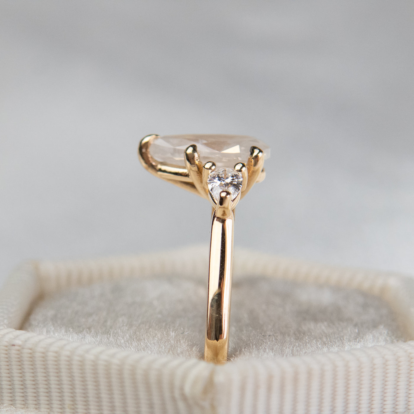 A side profile view of a three stone ring with a central pear-shaped diamond set in gold , flanked by two smaller pear diamonds, reflecting light on a soft, light, blurred background. 