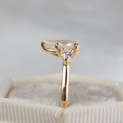 A side profile view of a three stone ring with a central pear-shaped diamond set in gold , flanked by two smaller pear diamonds, reflecting light on a soft, light, blurred background. 