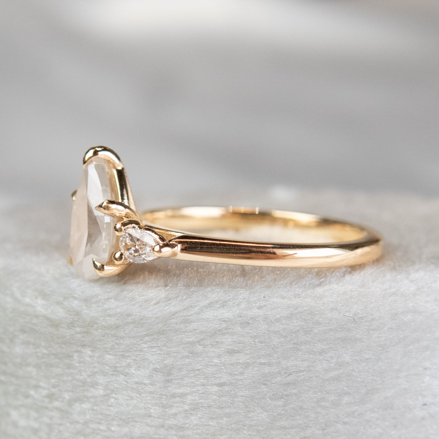 A three stone ring with a central pear-shaped diamond set in gold , flanked by two smaller pear diamonds, reflecting light on a soft, light, blurred background. 