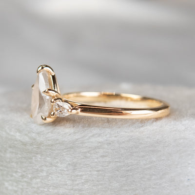 A three stone ring with a central pear-shaped diamond set in gold , flanked by two smaller pear diamonds, reflecting light on a soft, light, blurred background. 