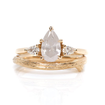 A three stone ring with a central fancy white pear-shaped diamond set in gold , flanked by two smaller pear diamonds, paired with an organic textured band. Displayed on a white background. 