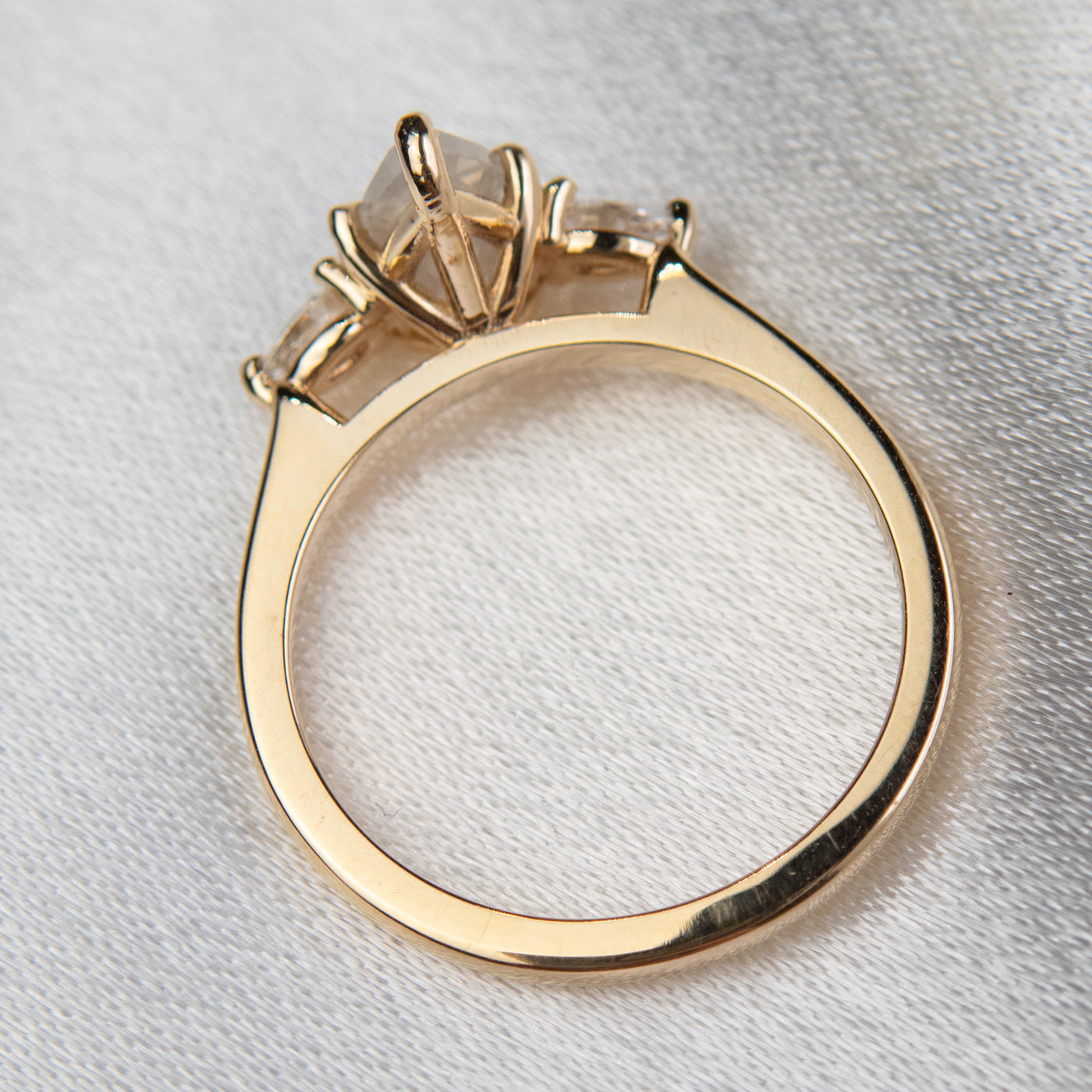 A three stone ring with a central pear-shaped diamond set in gold , flanked by two smaller pear diamonds, reflecting light on a soft, light, blurred background. 